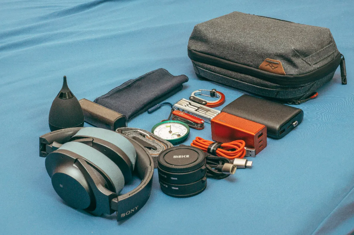 Travel gear laid out on a blue surface: a packing cube, wallet, headphones, camera and watch